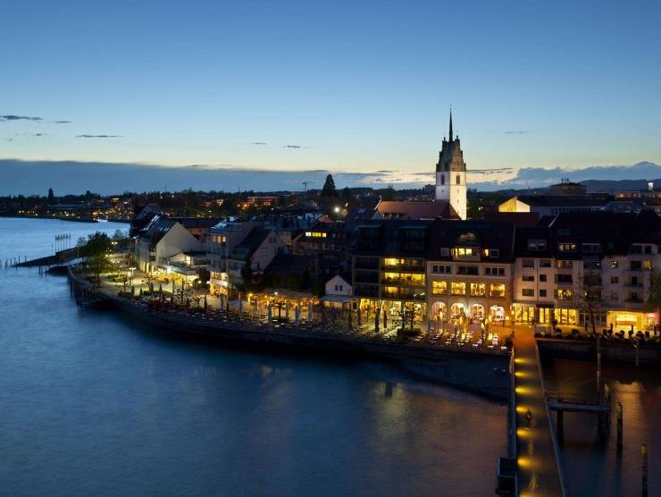 Luftaufnahme der Stadt Friedrichshafen bei Nacht mit beleuchteten Gebäuden am Bodenseeufer.